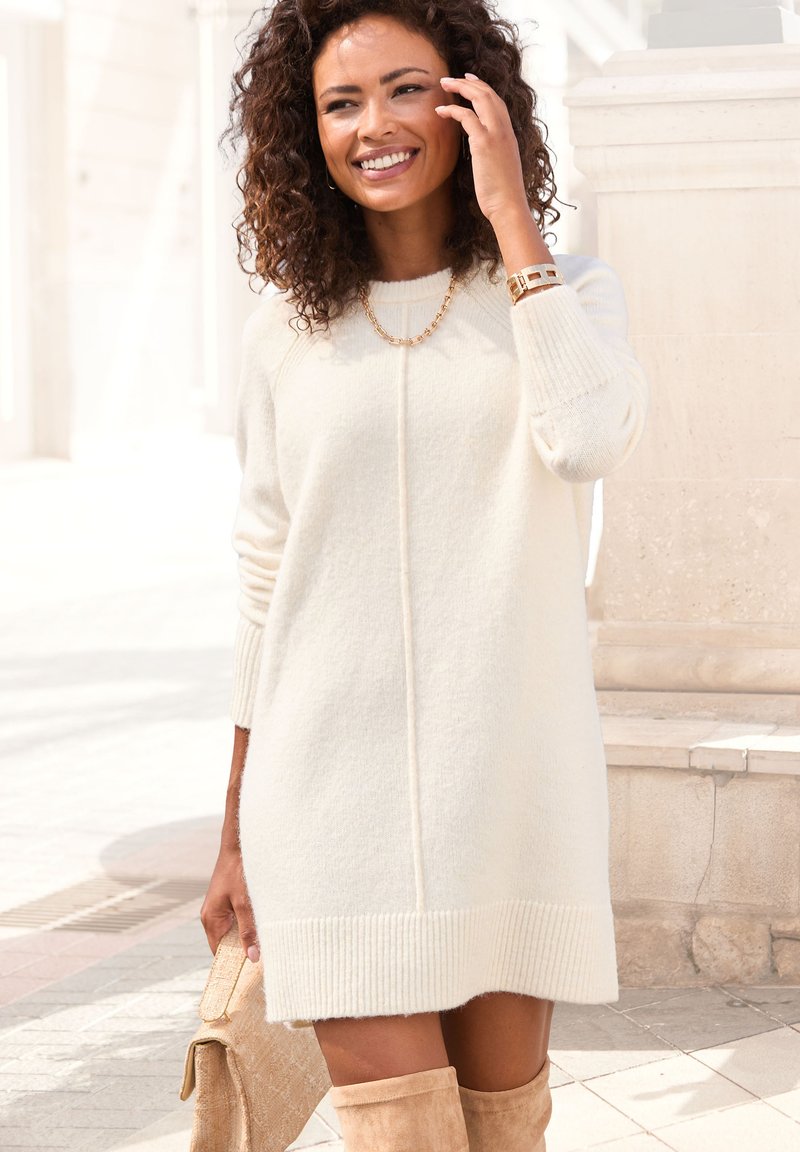 Strickkleid in Creme mit gerippten Bündchen und Saum, das mit einer zentralen Naht versehen ist. Kombiniert mit beigen kniehohen Stiefeln und einer beigen Tasche.