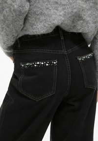 Crne traperice od denima s dva stražnja džepa, s geometrijskim ukrasima i srebrnim detaljima. Tkanina izgleda teksturirano i izdržljivo.