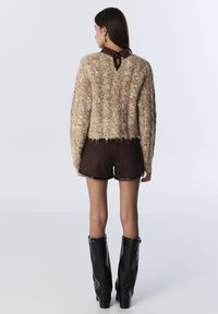 Suéter beige esponjoso recortado con detalle de ojal en la parte trasera, combinado con pantalones cortos de ante marrón y botas negras de tacón alto hasta la rodilla. Textura suave.