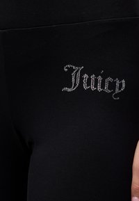 Μαύρο ύφασμα με τη λέξη "Juicy" γραμμένη με μικρές ασημένιες στρας στην περιοχή του άνω μηρού του ρούχου.