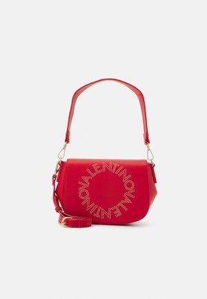 Sac à bandoulière en cuir rouge avec une forme incurvée, arborant un logo de marque circulaire en métal clouté. Sangle ajustable et quincaillerie en ton doré.