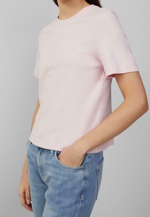 Person trägt ein hellrosa kurzärmeliges Hemd und blaue Jeans mit einer Hand in der Tasche, steht vor einem einfarbigen grauen Hintergrund.