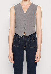 Ärmlös grå väst med fyra knappar, ihop med mörkblå jeans som har framfickor med klaff och kontrasterande sömmar.