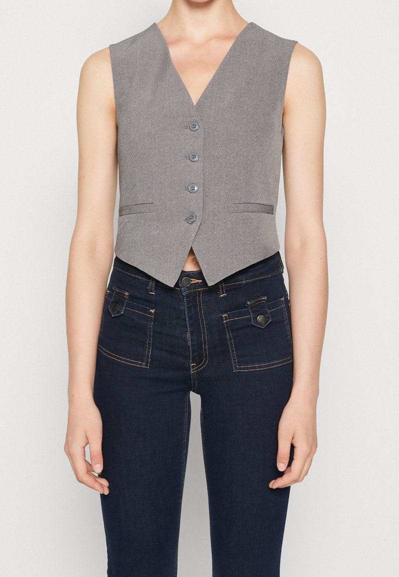 Ärmlös grå väst med fyra knappar, ihop med mörkblå jeans som har framfickor med klaff och kontrasterande sömmar.