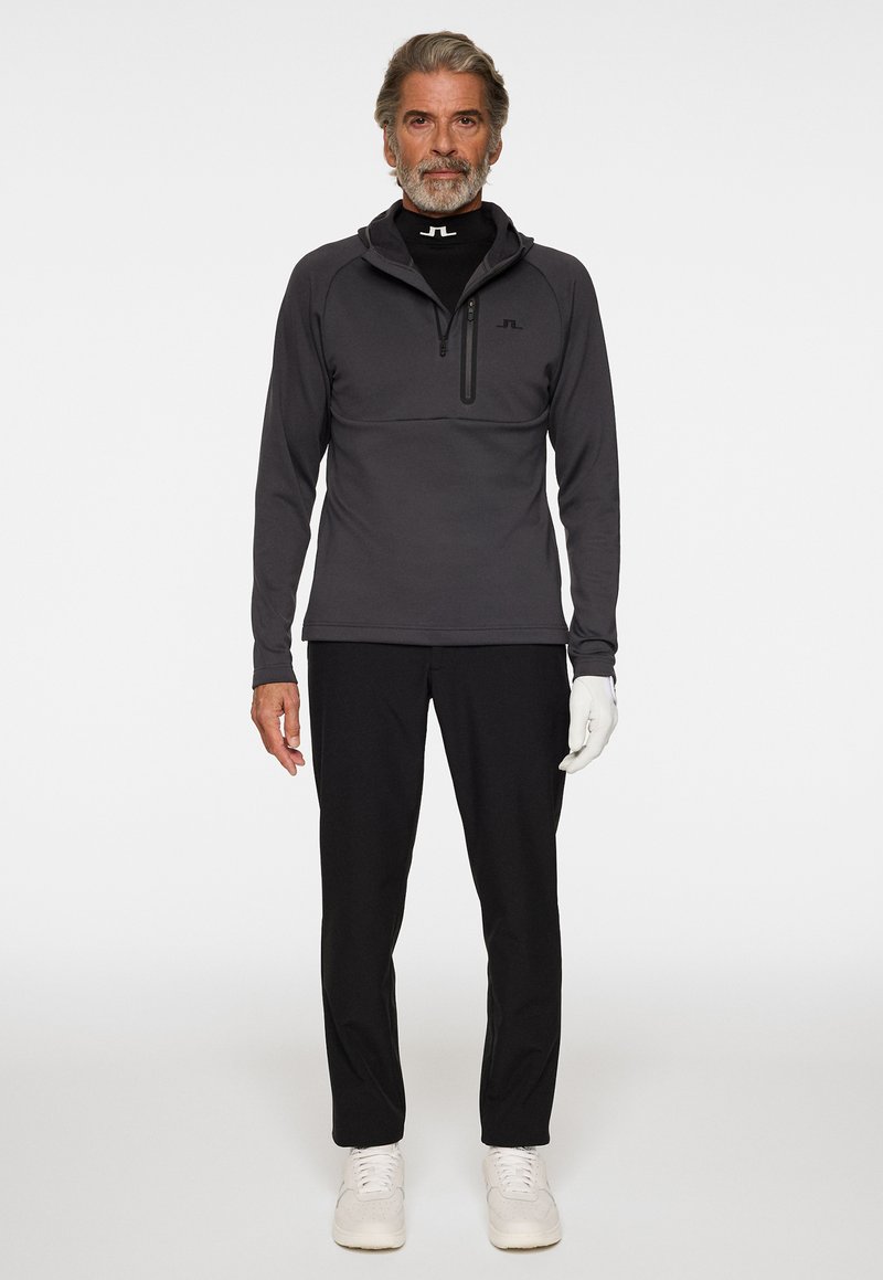 Grauer Reißverschluss-Pullover mit hohem Kragen, schwarzem Base Layer darunter, schwarzen Hosen und weißen Schuhen, mit einem dezenten Markenlogo auf dem Pullover.