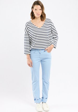 Femme debout portant un haut à manches longues rayé blanc et bleu marine, un pantalon bleu clair à revers et des baskets blanches, sur un fond blanc.