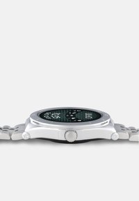 Reloj de plata redondo con una esfera negra con patrón, marcadores de hora blancos y una correa de plata texturizada. Presenta una corona en el lateral.