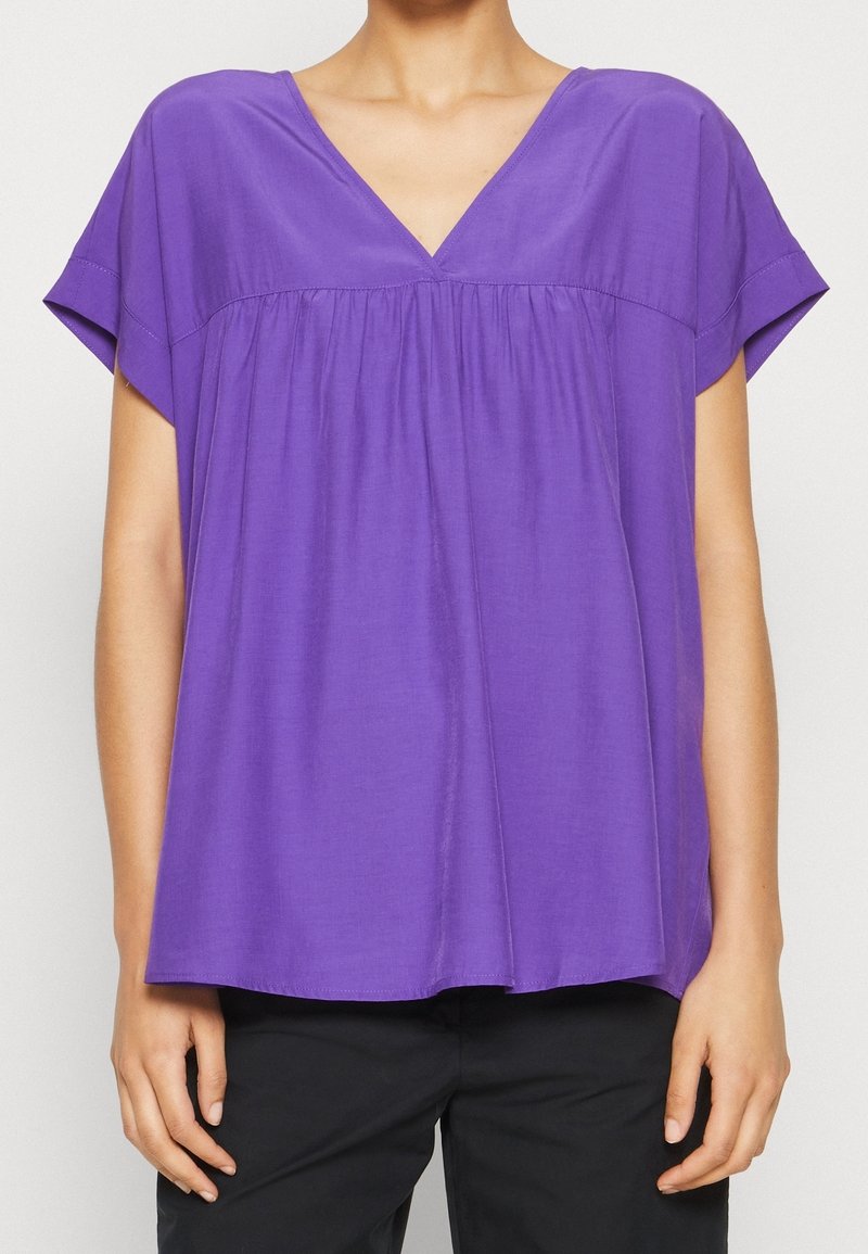 V-ringad lila blus med korta ärmar och samlad detalj fram. Tillverkad av mjukt, lätt tyg.