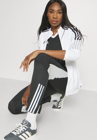 Witte zip-up jack met zwarte strepen, zwarte legging met witte accenten en grijze sportschoenen met witte strepen. Alle items zijn sportkleding.
