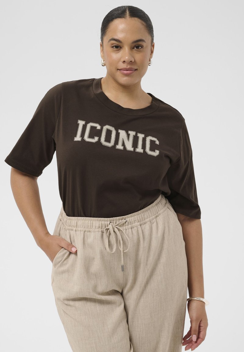 Femme aux cheveux tirés en arrière, portant un t-shirt noir « ICONIC » et un pantalon beige à cordon, debout avec une main dans la poche.