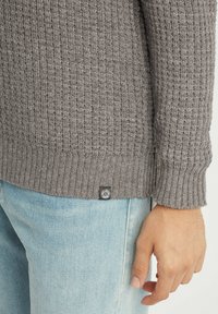 Grauer Strickpullover mit gerippter Textur und subtilen Mustern, seitlichem Schlitz und schwarzem Logo-Tag nahe dem Saum, kombiniert mit hellblauen Jeans.