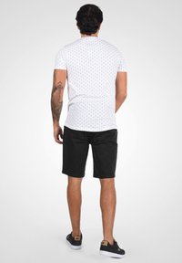 Weißes Kurzarmhemd mit kleinen schwarzen Punkten, kombiniert mit schwarzen Shorts. Das Model steht mit dem Rücken zur Kamera und zeigt ein Tattoo am Arm.