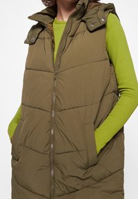 Olejozelená péřová vesta se zapínáním na zip, vysokým límcem a ramenními popruhy na cvoky; má boční kapsy a prošívaný vzor.