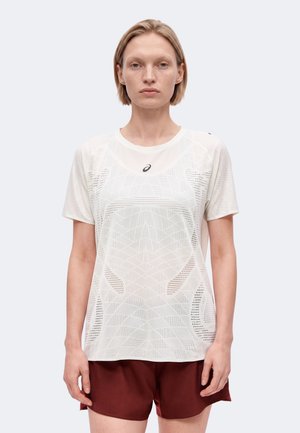 Frau trägt ein weißes, gemustertes Sport-T-Shirt mit kurzen Ärmeln und dunkelroten Shorts und steht vor einem einfarbigen hellen Hintergrund.