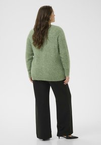 Pull en tricot vert à la texture douce et manches longues, accompagné d'un pantalon noir à jambes larges et de chaussures à talons bas, vu de dos.