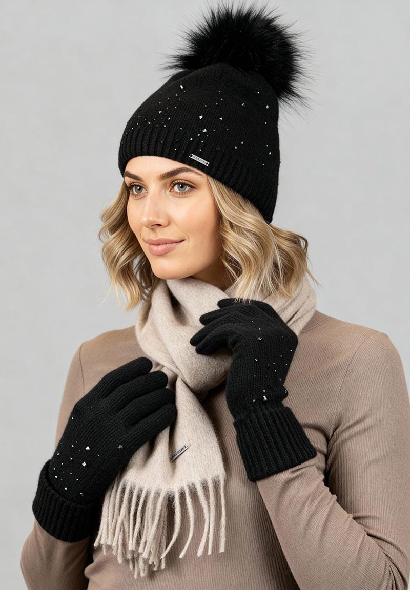 Zwarte gebreide muts met een pompon, bijpassende zwarte handschoenen met kleine versieringen en een lichtbeige sjaal met franjes.