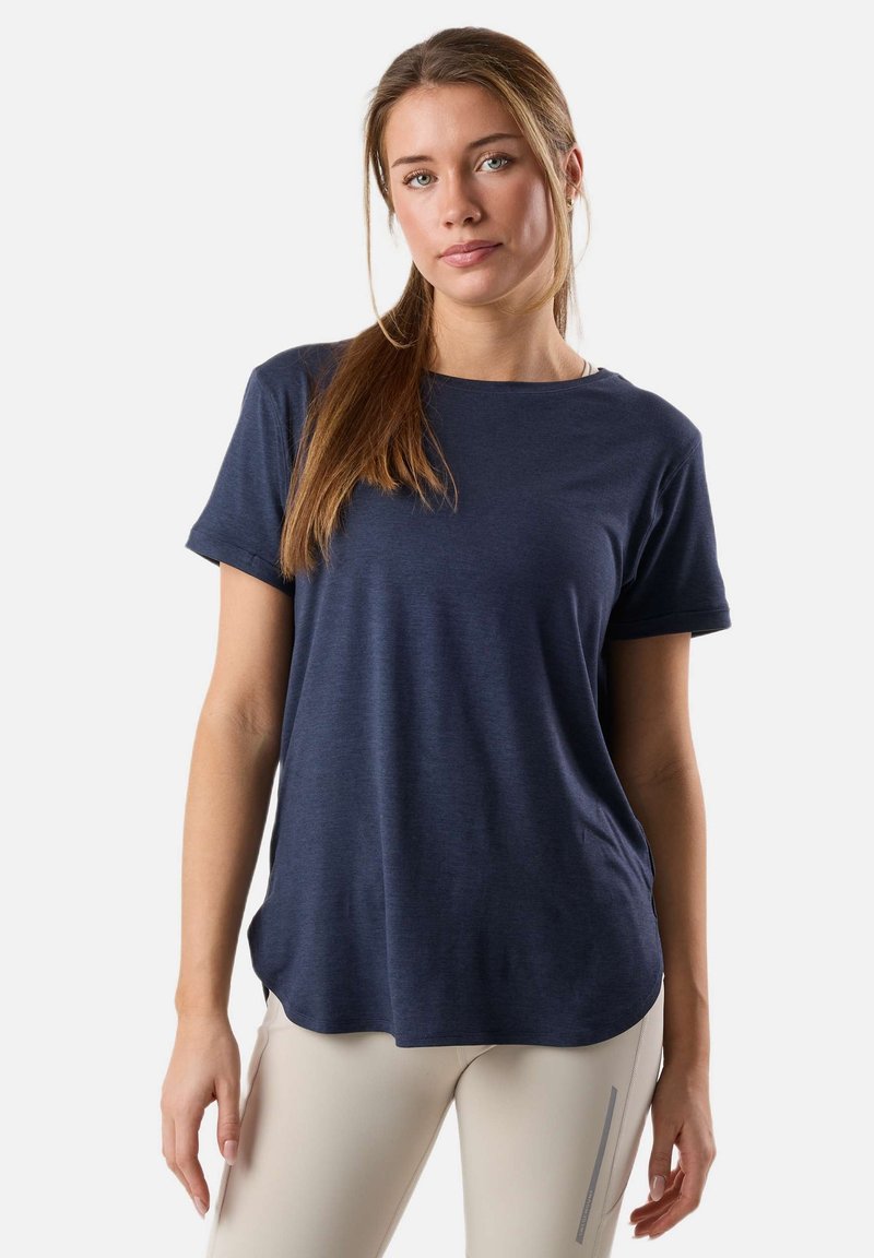 Young woman with long brown hair in a ponytail wearing a navy blue t-shirt and light beige pants against a plain light background.