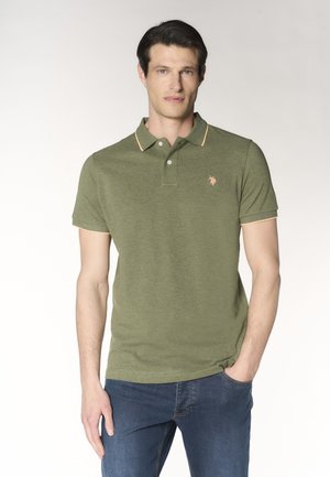 Uomo con capelli scuri che indossa una polo verde oliva con bordi arancioni e jeans blu, in piedi con una mano in tasca su sfondo bianco.