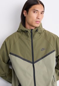 Olivengrøn Nike-jakke med hætte, lynlåslukning og kontrasterende grå og lysere grønne diagonale paneler. Tekstureret stof med synlig syning.