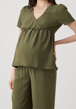 Femme portant un haut de maternité ample vert olive et un pantalon assorti, debout avec une main sur la hanche.