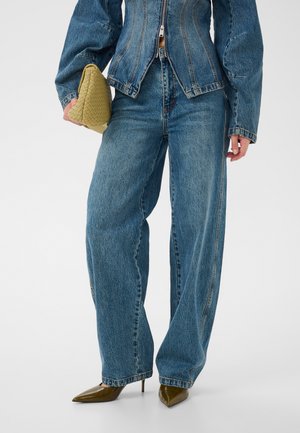 Frau trägt weite blaue Jeans und eine taillierte Jeansjacke, hält eine texturierte beige Clutch und trägt spitze braune High Heels.
