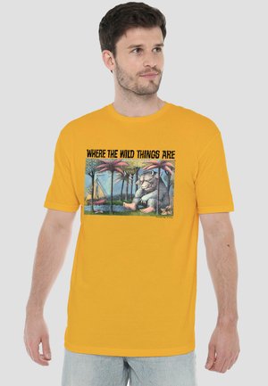 Mann trägt gelbes T-Shirt mit dem Aufdruck „Where the Wild Things Are“ und einer Illustration eines Wesens, das neben Palmen und einem Segelboot sitzt.