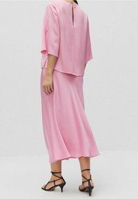 Nainen, joka on pukeutunut löysään vaaleanpunaiseen paitapuseroon ja väriltään matching virtaavaan midi-hameeseen, joissa on mustat nyörisaappaat, seisoo yksinkertaista harmaata taustaa vasten.