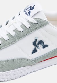 Zapatilla con upper de textil blanco y acentos de ante gris, suela de goma y un emblema azul en el lateral y en la lengüeta.