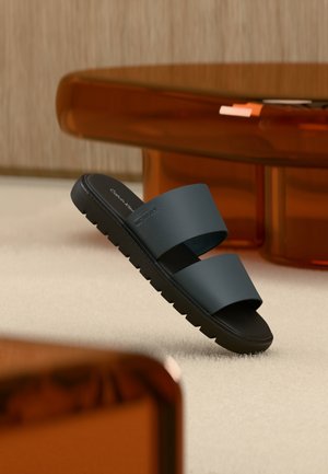 Sort slidesandal med to brede remme, serrateret sål og Calvin Klein-logo, placeret opret på beige tæppe nær et brunt bord.