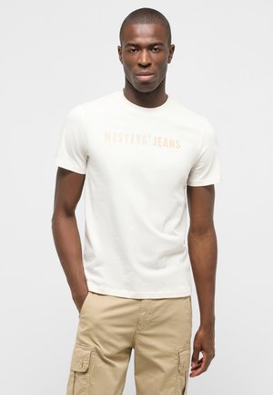 Junger Mann mit dunkler Haut, der ein weißes "Mustang Jeans" T-Shirt und beige Cargohosen trägt, steht mit einer Hand in der Tasche vor weißem Hintergrund.