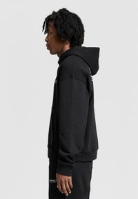 Czarny hoodie wykonany z miękkiego materiału, z przednią kieszenią, prążkowanymi mankietami i kapturem. Projekt zawiera minimalne logo z przodu.