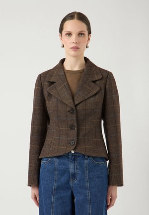 Donna che indossa un blazer marrone a quadri con tre bottoni sopra una maglia marrone e jeans blu a vita alta, in piedi davanti a uno sfondo chiaro e uniforme.