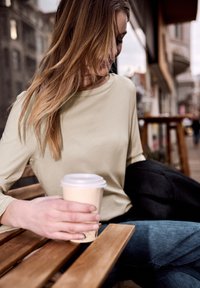 Jeune femme assise à une table de café en plein air, tenant une tasse de café à emporter, portant un haut beige et un jean bleu, avec un décor urbain en arrière-plan.