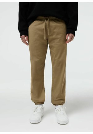 Osoba ubrana w beżowe spodnie z sznurkiem w pasie i elastycznymi mankietami oraz białe trampki, stojąca na jednolitym jasnym tle.