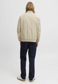 Veste bomber beige avec taille élastique et poignets, associée à un jean droit bleu marine et des baskets blanches. Vue arrière simple.