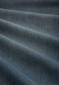 Denim stof in donkere blauwtinten, met een gestructureerd oppervlak en duidelijke diagonale weefpatronen, gelegd in zachte, vloeiende plooien.