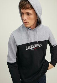 Mikina s kapucňou v dvoch farbách, šedej a čiernej, vyrobená z jemnej látky, s logom potlačeným na hrudi.