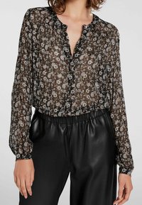 Zwarte bloemenblouse gemaakt van doorzichtig materiaal met een gerimpelde kraag, knoopsluiting aan de voorkant en lange pofmouwen, gecombineerd met zwarte broek met hoge taille.