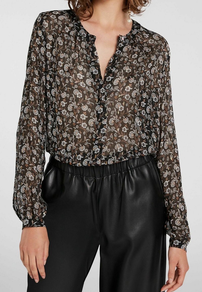Zwarte bloemenblouse gemaakt van doorzichtig materiaal met een gerimpelde kraag, knoopsluiting aan de voorkant en lange pofmouwen, gecombineerd met zwarte broek met hoge taille.