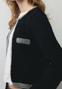 Mujer vestida con un cárdigan de punto negro con detalles plateados y bolsillos, sobre una camisa blanca, mostrada desde los hombros hasta la cintura.