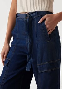 Pantaloni in denim blu scuro con vita alta, passanti per cintura, cintura annodata e tasche laterali con cuciture visibili e una vestibilità ampia.