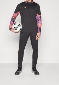 Puma Sportbekleidungsset mit einem schwarzen eng anliegenden Oberteil mit bunten gemusterten Ärmeln, schwarzen Hosen und schwarzen Sportschuhen, während er einen Fußball hält.