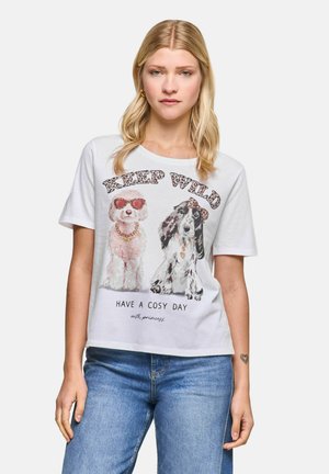Woman wearing a white T-shirt with illustrations of two dogs, decorative text "Keep Wild," and the message "Have a cosy day." Blue jeans.