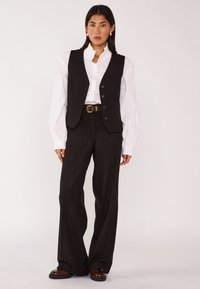 Zwarte vest over een witte shirt, met een hoge kraag en knoopsluiting. Wijde zwarte broek met een bruine riem en bruine schoenen.