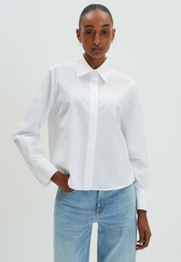 White long-sleeve collared shirt in smooth, lightweight fabric with a button-down front and a cropped hem. Worn with light blue denim jeans.