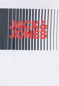 Balta T-kreklā ir drosmīgs grafisks attēls ar "JACK & JONES" spilgti sarkanā krāsā uz plānu melnu vertikālu svītru fona.