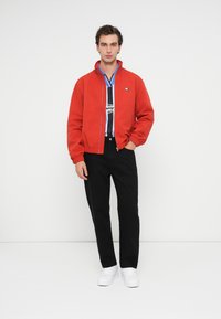 Κόκκινη bomber τζάκετ με ελαστικούς μανικιών, με μπλε αντίθεση στη φόδρα, φορεμένη πάνω από ένα ασπρόμαυρο ριγέ πουκάμισο, συνδυασμένη με μαύρο παντελόνι και λευκά αθλητικά παπούτσια.