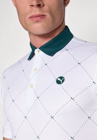 Hvid polo-shirt med en mørkegrøn krave, der har et diamantmønster og et lille grønt Puma-logo på brystet. Knaplukning ved halsen.