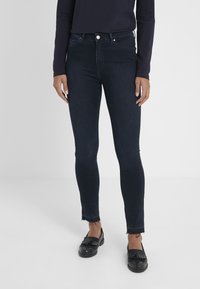 Jeans skinny de color azul oscuro con una cintura alta, presentan un dobladillo sin acabar y un ligero desgastado. Combinados con unos mocasines negros y una blusa de mangas largas.