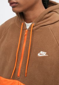 Hnedá fleecová mikina s oranžovým zipsom a šnúrkami, na hrudi s bielym logom Nike a hladkým, textúrovaným materiálom.